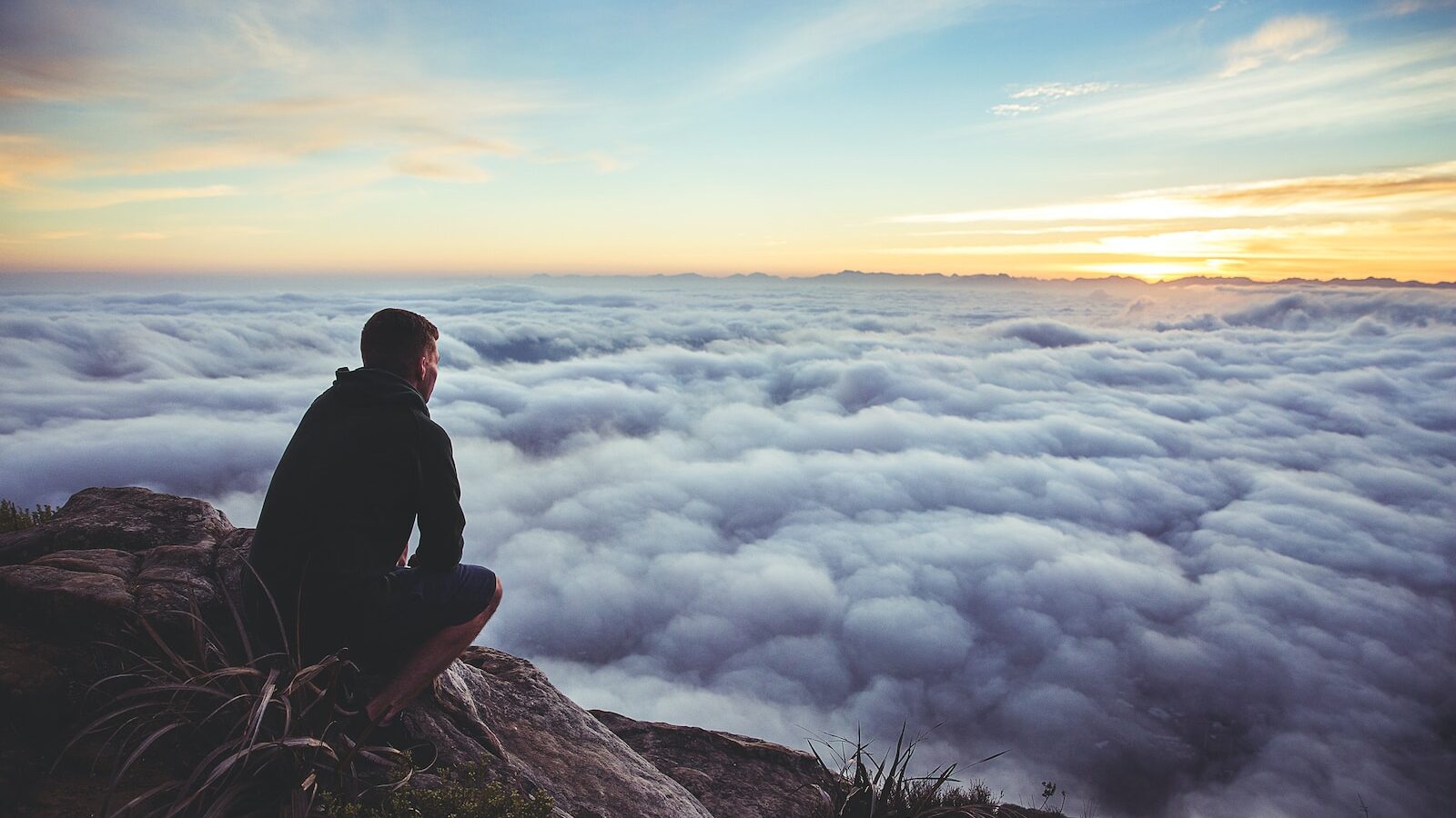 man sitting on gray rock while staring at white clouds