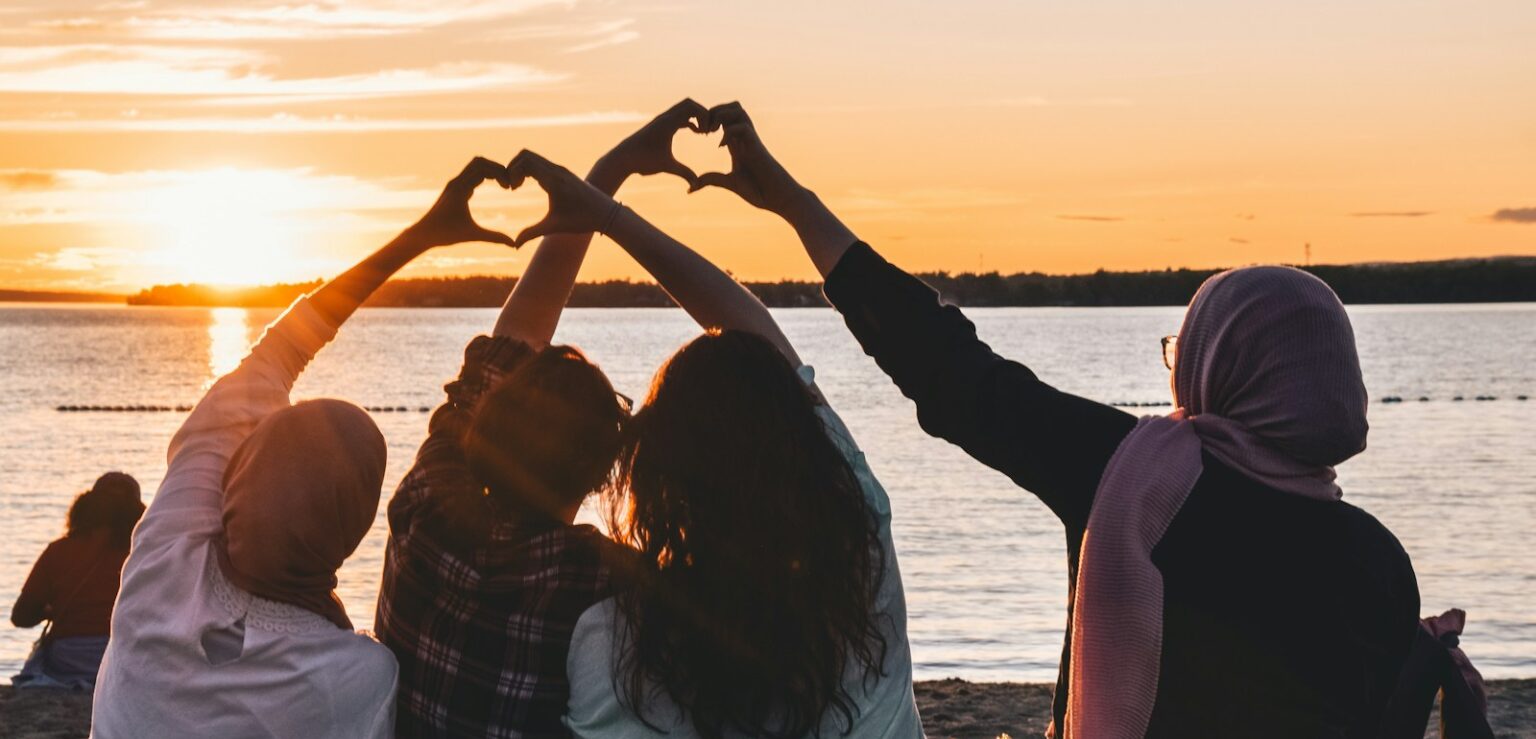 four people sitting on shore forming hearts with their hands during golden hour